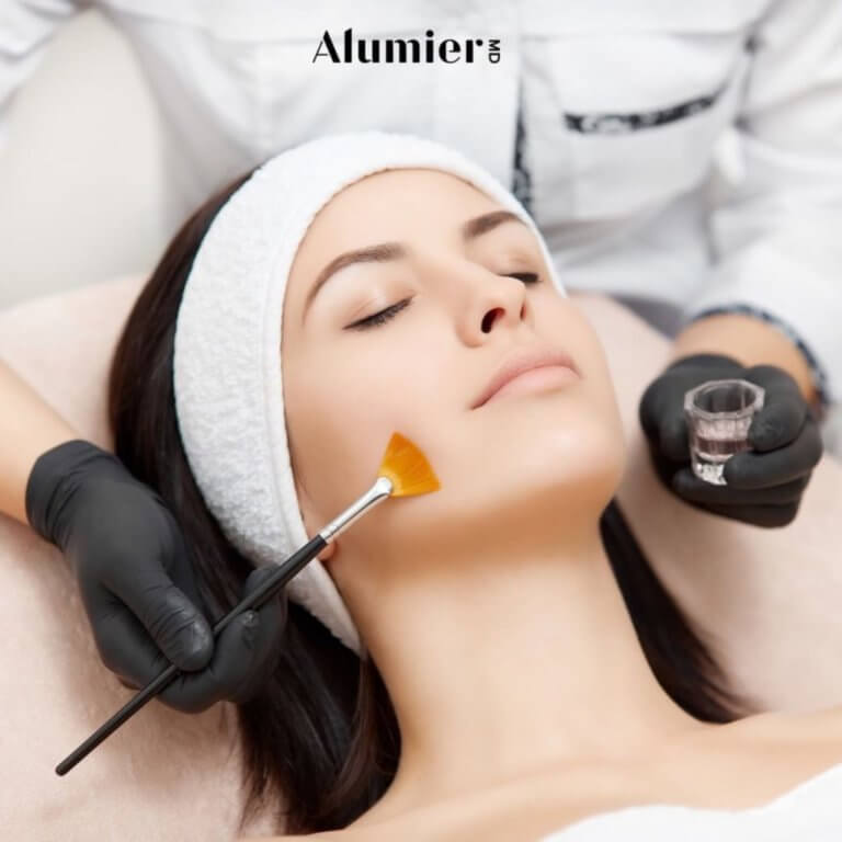 Picture of a woman laying face up receiving an paint brush stroke of AlumnierMD branded Chemical Peel